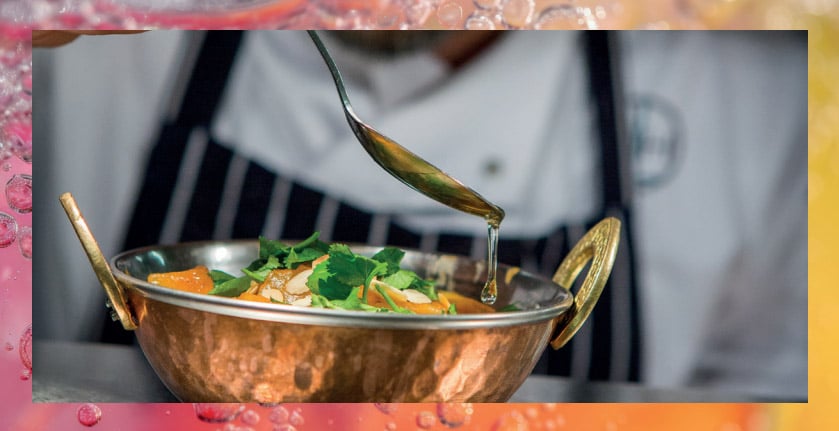 A chef drizzles sauce over a vibrant dish in a decorative copper bowl, garnished with greens and citrus slices.